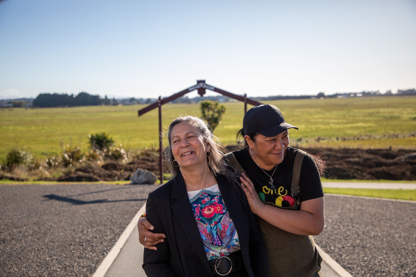 Two wāhine laughing arm in arm on the path, with the waharoa and farmland behind