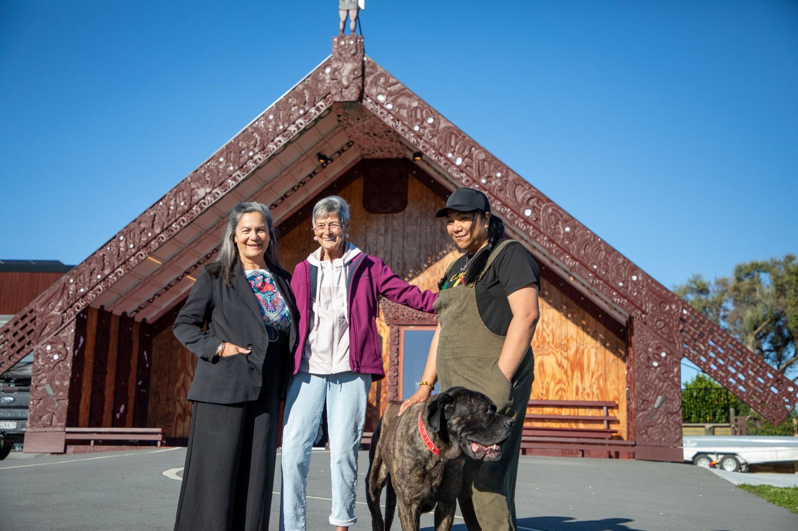 Three wāhine and a dog in front of the carved wharenui